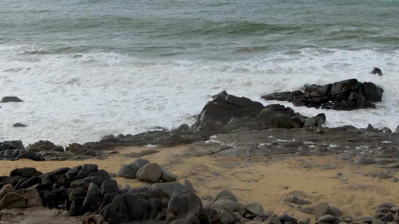 Foam washes over flat shoreline rocks below a gray-green ocean band at Punta Gorda, with tan sand visible along the lower frame and no visible people.
