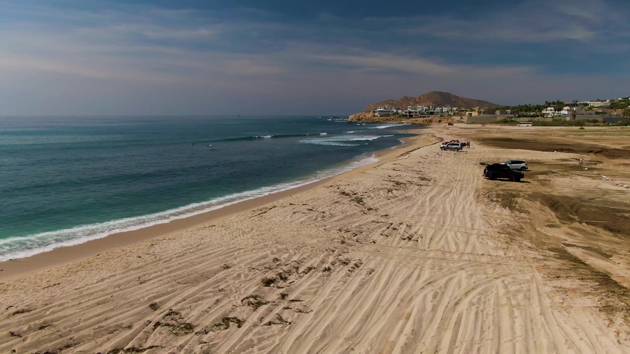 Aerial view over El Tule Los Cabos with turquoise ocean on the left, a tire-marked sandy beach on the right, vehicles near the shoreline, distant buildings, a rocky hill, and no visible people.