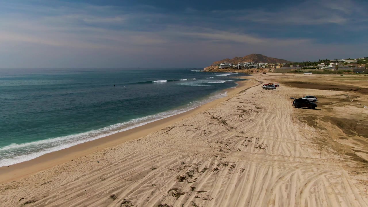 Drone view at El Tule Los Cabos with the foamy wave edge lower on the beach, vehicles farther along the sand, dry tire tracks across the foreground, and no visible people.