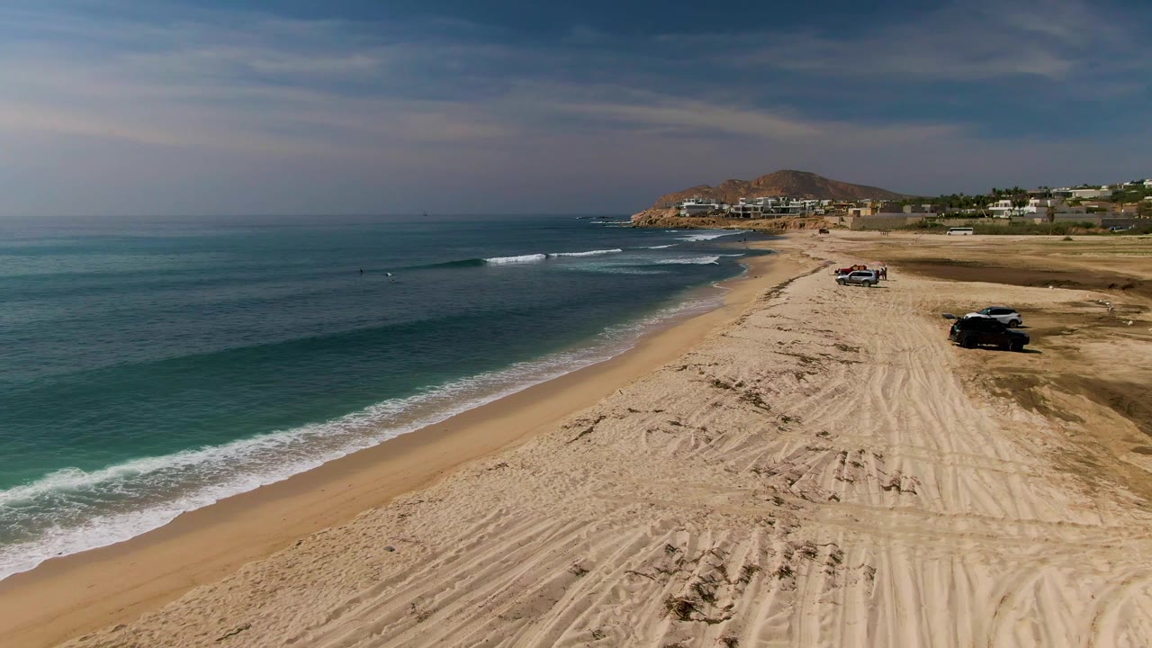High coastal view in El Tule Los Cabos with a wider band of turquoise water, white foam tracing the shore, vehicles along the right side of the beach, distant development, and no visible people.