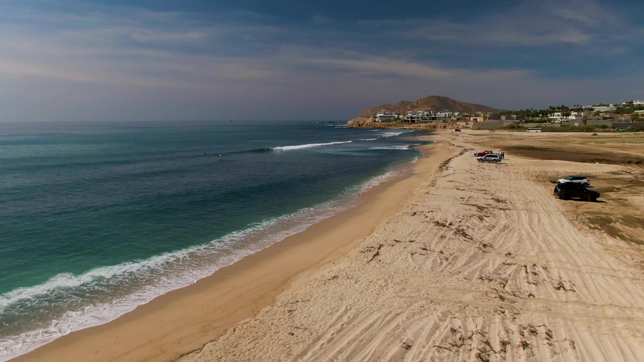 Aerial beach scene with ocean surf, empty sand, tire tracks, and parked vehicles.