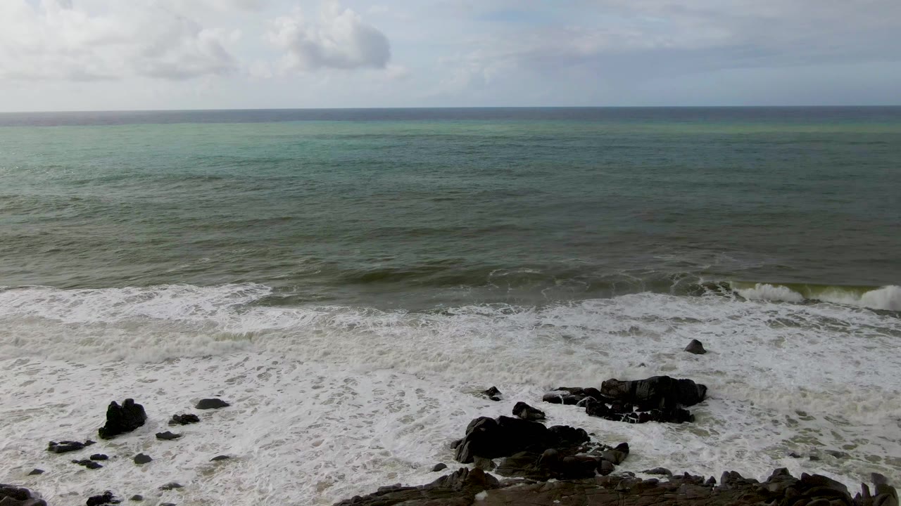 At Punta Gorda in Baja California Sur, white surf spreads across dark rocks below a turquoise-green ocean and gray sky, with no visible people.
