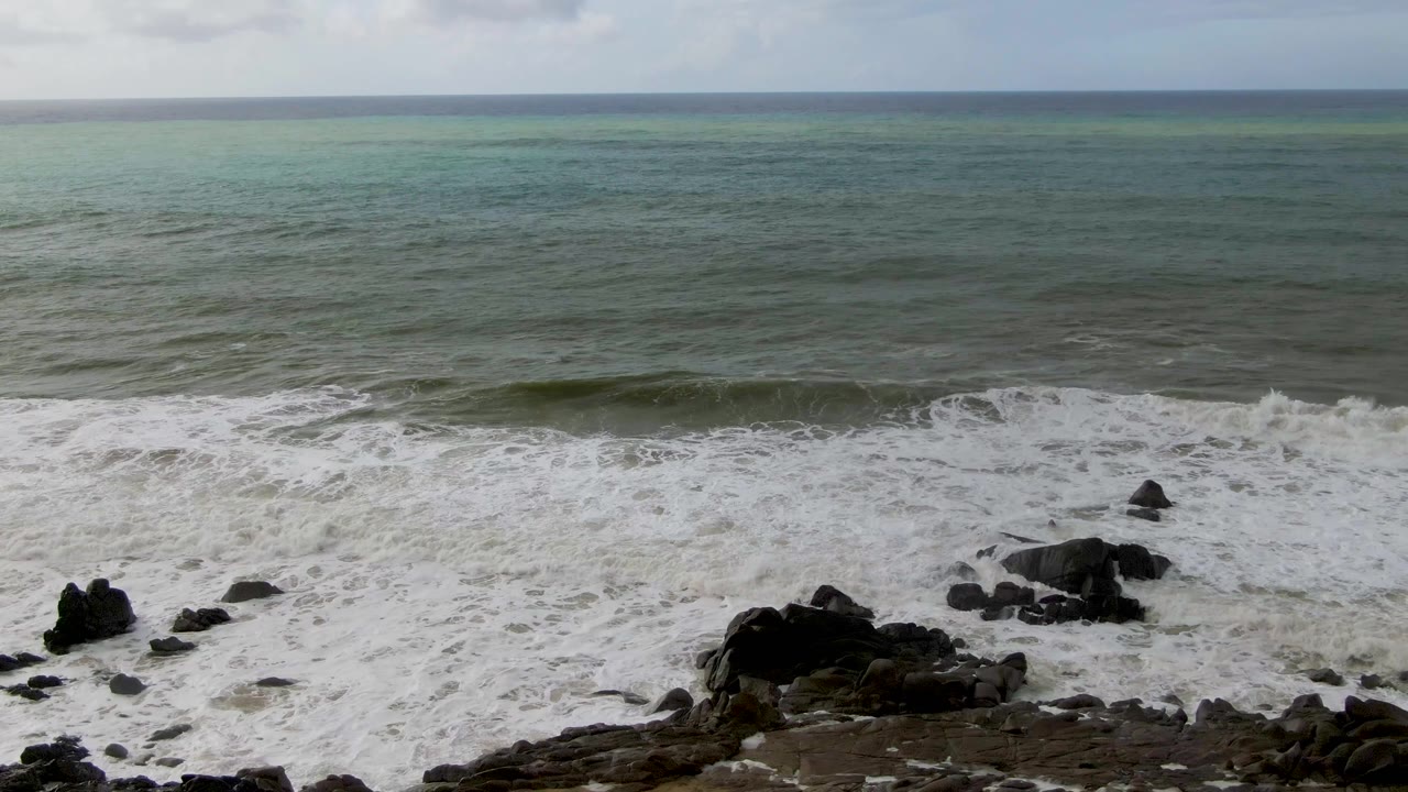 Whitewater collapses across central dark rocks on the Punta Gorda shoreline while muted green ocean fills the background and no people are visible.