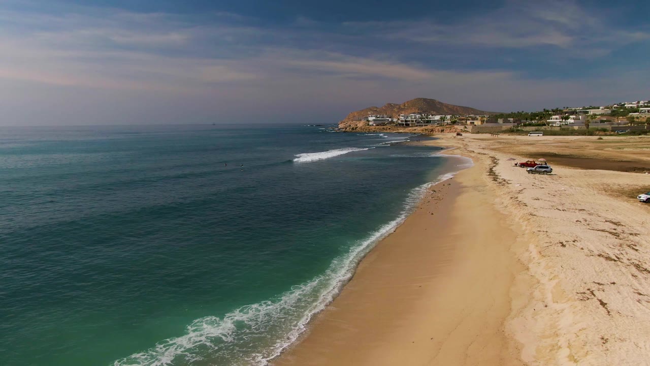 Aerial frame from El Tule Los Cabos showing blue-green ocean on the left, a sandy beach on the right, white surf at the shoreline, a rocky hill and buildings in the background, and no visible people.
