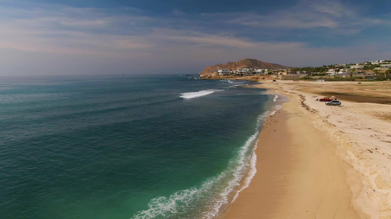 Aerial frame from El Tule Los Cabos with a longer white wave line reaching the sand, dark blue water offshore, turquoise water near shore, parked vehicles on the beach edge, and no visible people.