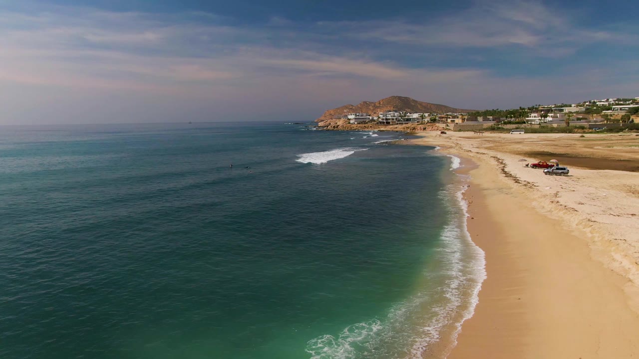 Aerial frame from El Tule Los Cabos showing the shoreline curve shifted farther right, small surf breaks offshore, parked vehicles near the upper beach, distant buildings, a rocky hill, and no visible people.