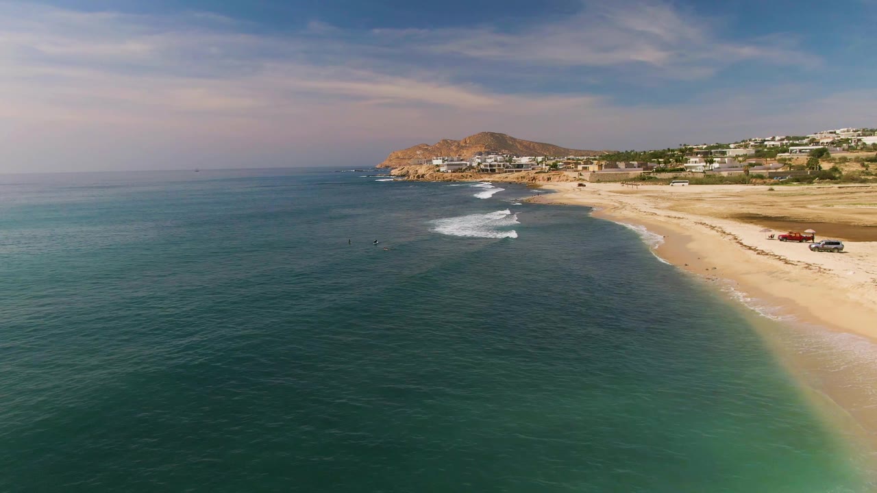 Aerial frame from El Tule Los Cabos showing turquoise water in the foreground, a sandy beach at right, distant coastal buildings, a low hill, and tiny swimmers offshore.