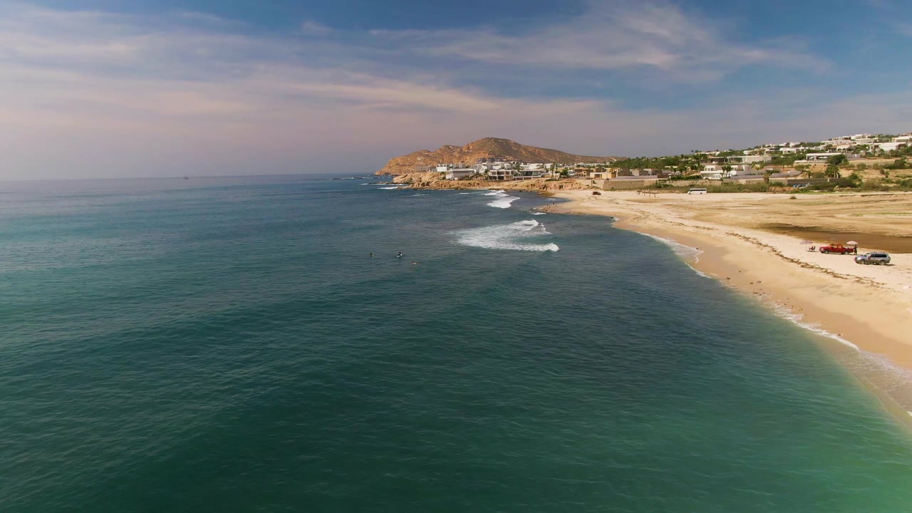 At 0:01, the El Tule Los Cabos shoreline remains angled across the frame with darker offshore water, small breaking waves, beach vehicles, buildings, and the hill beyond.