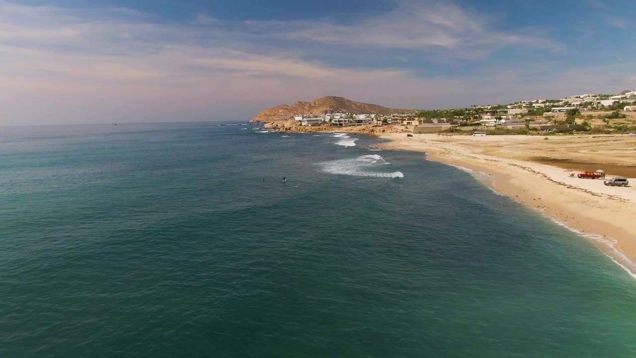 At 0:02, more open water dominates the foreground while the sandy El Tule Los Cabos beach curves toward coastal buildings and a low hill under streaked cloud cover.