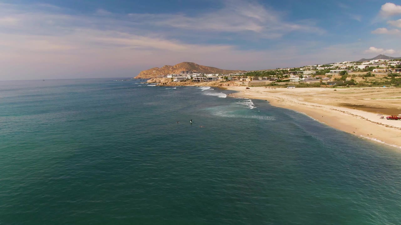 Aerial view at El Tule Los Cabos showing turquoise water in the foreground, a sandy beach curving toward a coastal hill, white buildings inland, and small people visible near the shore.