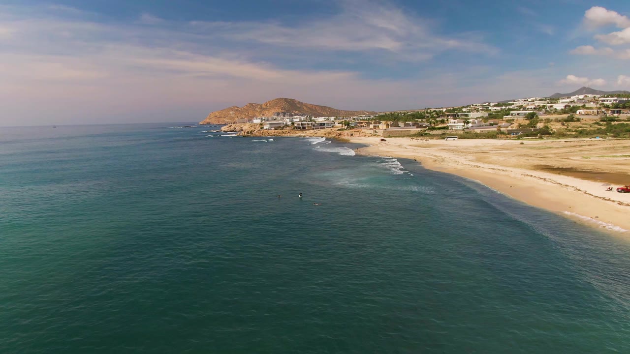 Wide aerial coastline with calm water, surf lines, beach, hills, and buildings.