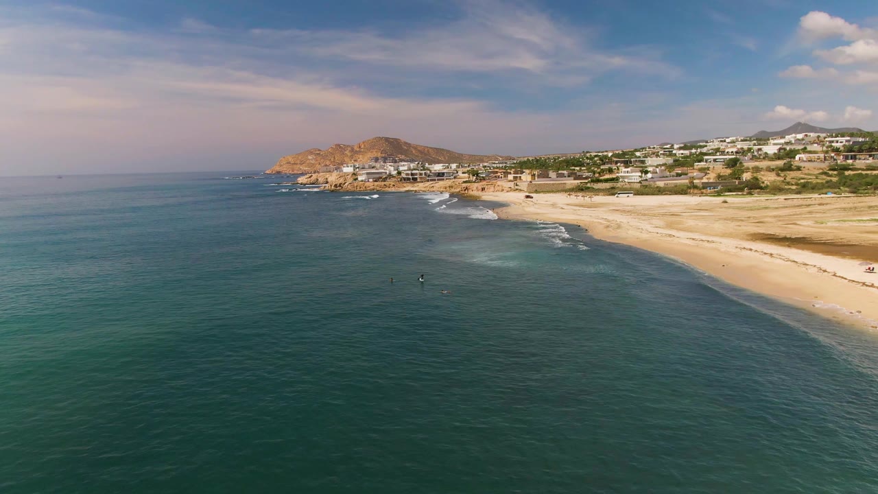 El Tule Los Cabos coastline seen from above, with open ocean in front, pale sand along the right side, small figures near the waterline, and a rocky hill with buildings in the distance.