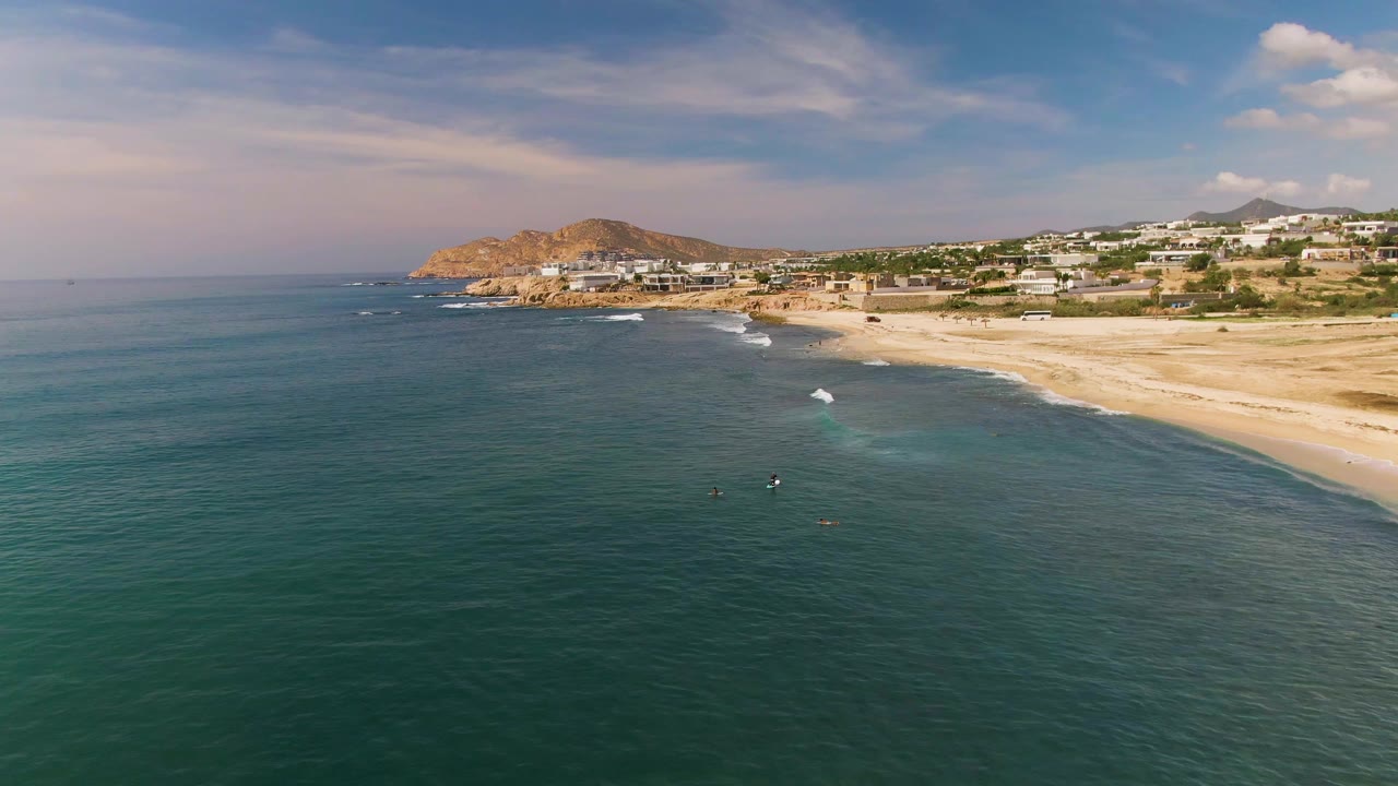 Aerial frame over El Tule Los Cabos with teal water in the foreground, small surfers near the break, a sandy beach, low buildings, dry hills, and blue sky.