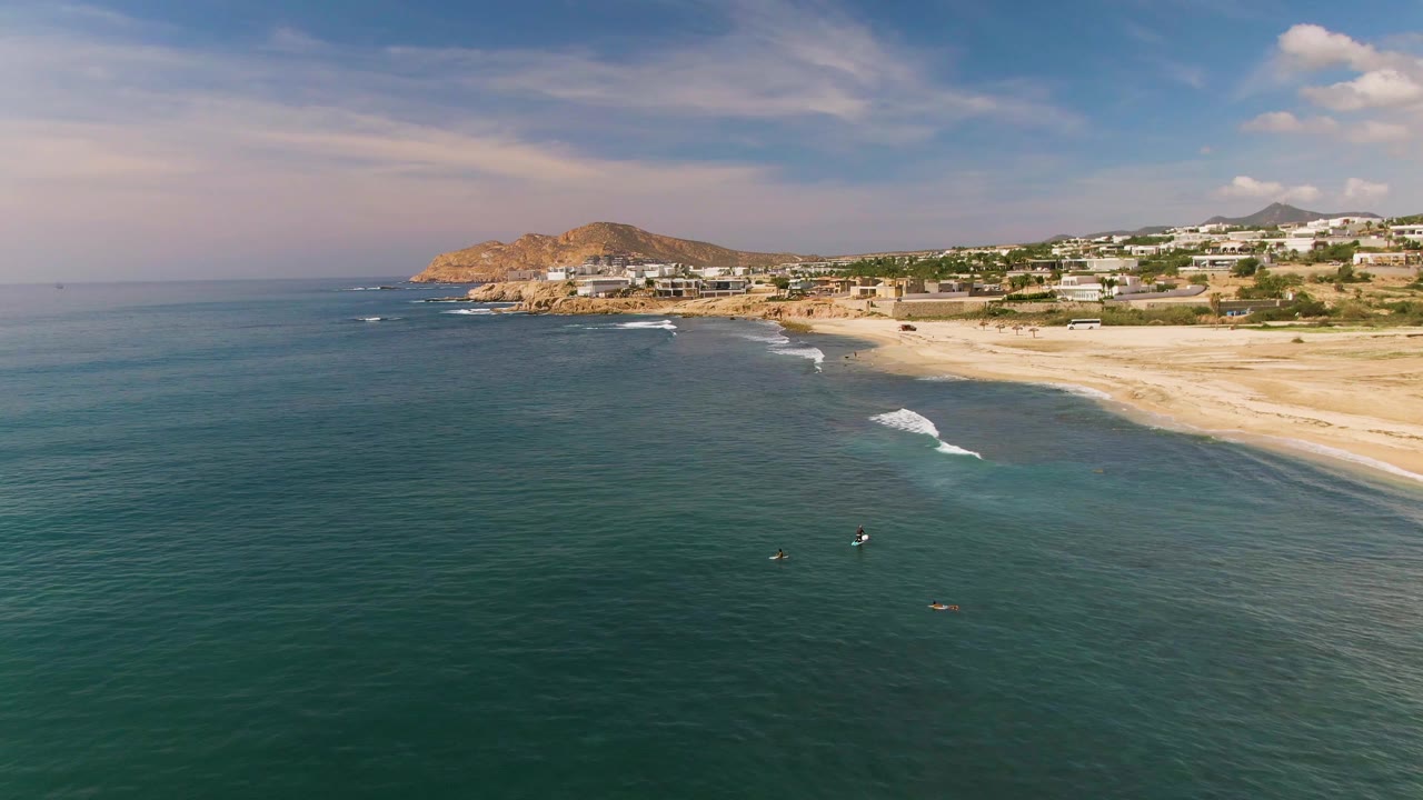 Aerial view at El Tule Los Cabos with more open water filling the foreground, small people in the surf zone, and a pale sandy beach curving below coastal buildings.