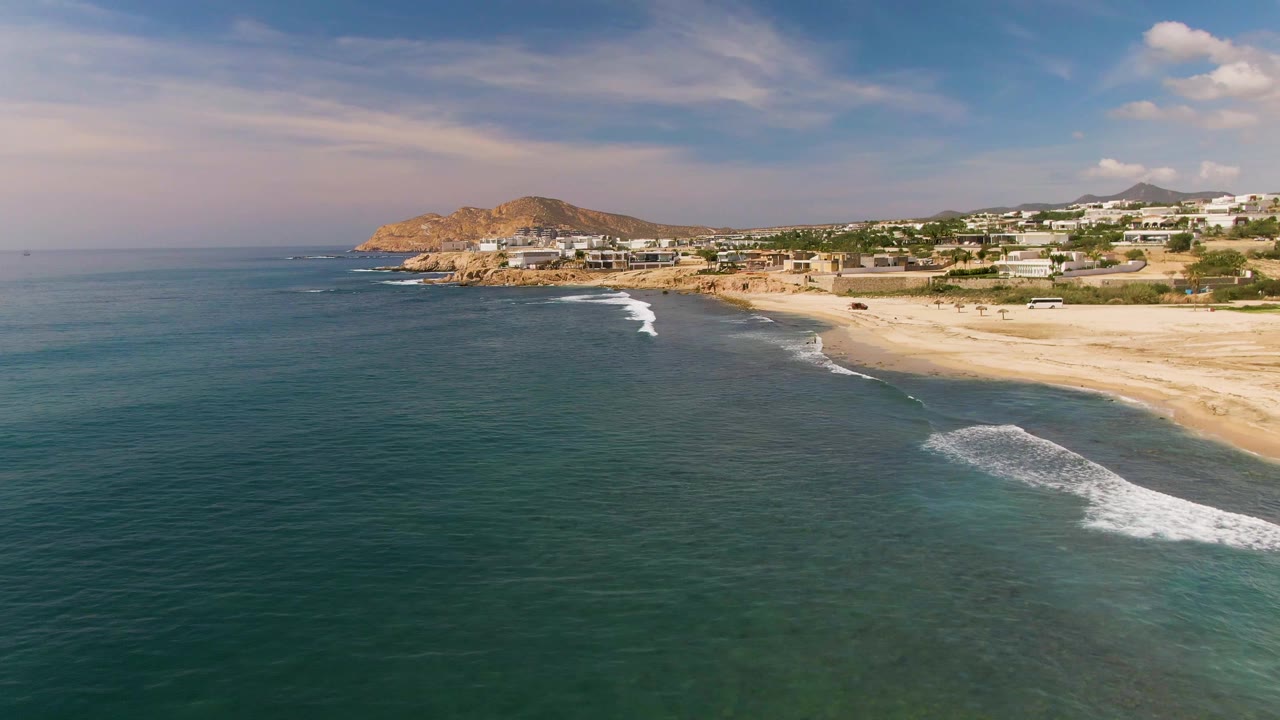 Opening frame over El Tule, Los Cabos with blue-green water in the foreground, a pale sandy beach to the right, hillside buildings in the distance, and no visible people.