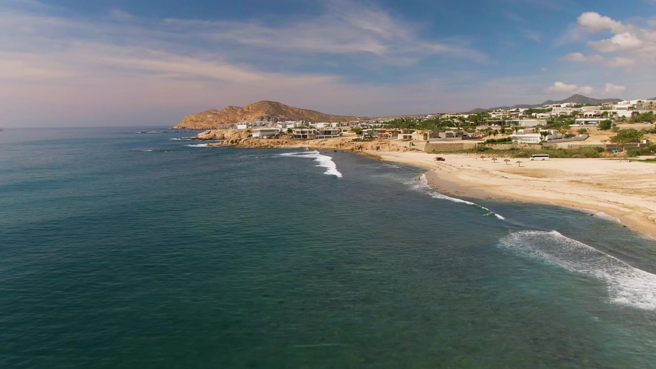 Blue ocean beside a sandy shore with white waves and hillside buildings.