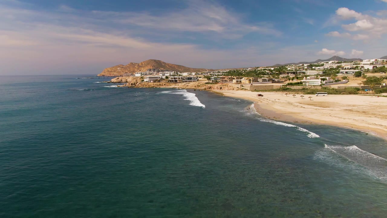 Aerial frame of El Tule with more open water in the foreground, a breaking wave near the lower right, the sandy beach curving along the coast, and no visible people.