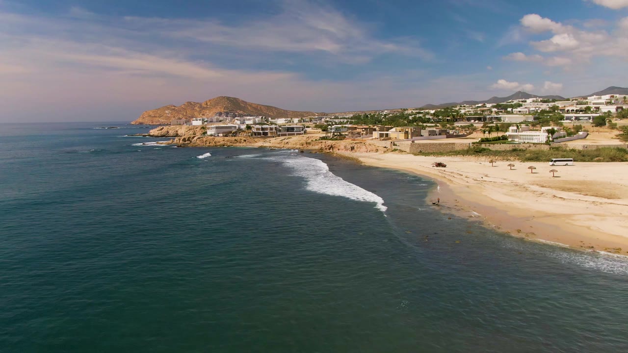 Aerial frame over El Tule Los Cabos with blue-green water, a curving white wave line, sandy shore, low buildings, rocky hill, and no visible people.