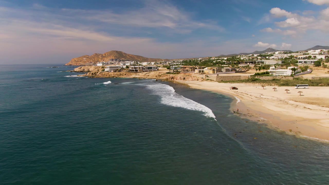 Aerial coastline view with a breaking wave moving toward a sandy beach and buildings beyond.