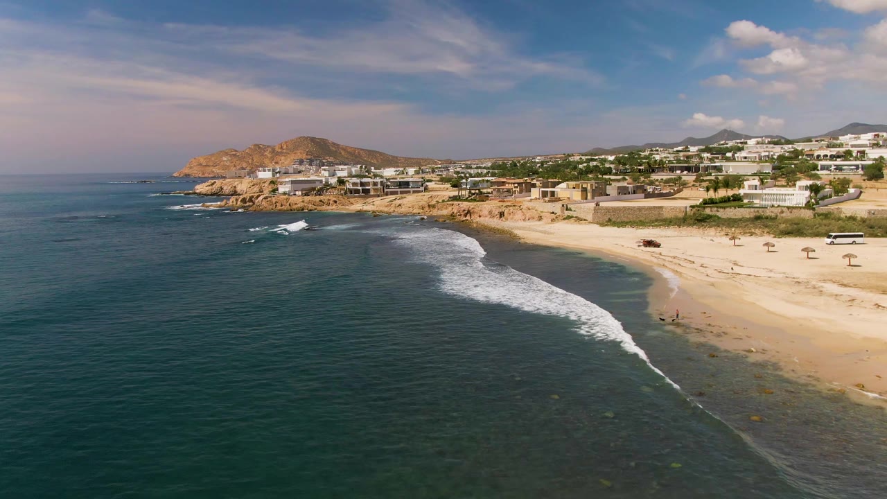 Aerial frame from El Tule Los Cabos showing a longer white surf band near the shoreline, dark blue-green water, pale sand, hillside buildings, and no visible people.