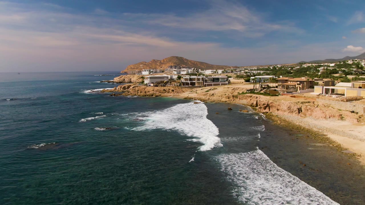 Aerial frame over El Tule Los Cabos with dark teal ocean in the foreground, white surf curling toward tan rocky cliffs, cliffside buildings, distant hills, and no visible people.