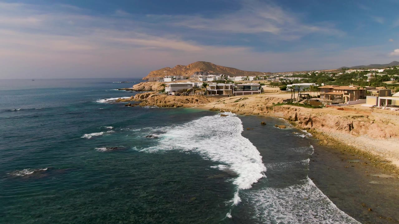 Foamy wave line advances toward a rocky beach below cliffside buildings.