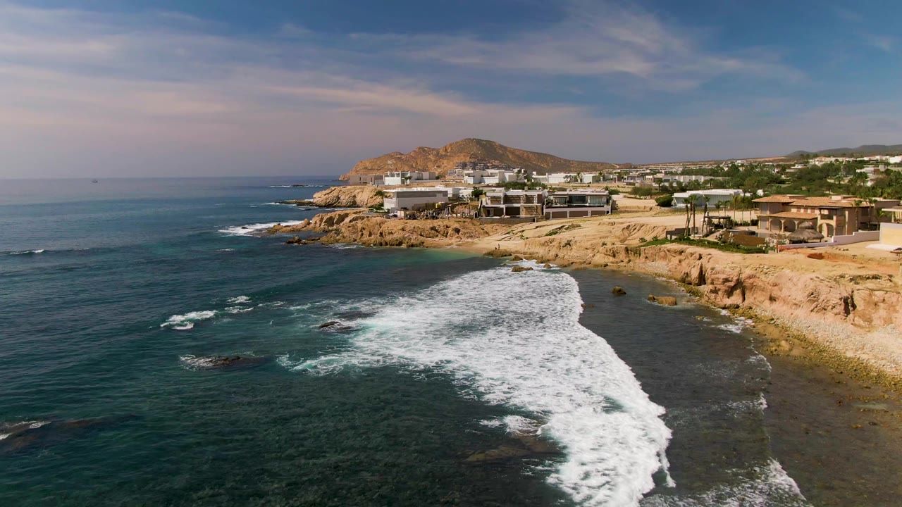 At two seconds, the breaking wave forms a broad curved band below the El Tule Los Cabos cliff, with scattered dark rocks in the water, hillside development beyond, and no visible people.