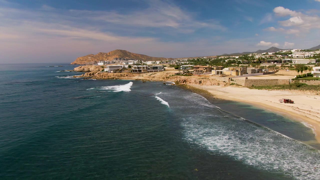 Aerial frame from El Tule Los Cabos showing dark blue-green ocean, white surf moving toward a sandy beach, rocky cliffs, low coastal buildings, and no visible people.