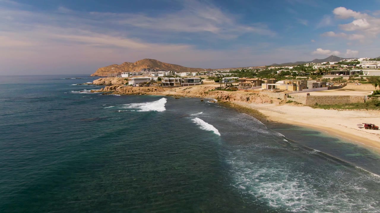 El Tule Los Cabos aerial view with a larger white wave line breaking near shore, turquoise shallows along the sand, rocky coastline, coastal buildings, and no visible people.