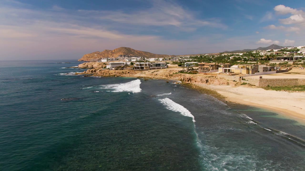 Aerial frame over El Tule Los Cabos with multiple white surf bands over dark reef-toned water, a pale sand beach at right, rocky cliffs and low buildings behind, and no visible people.