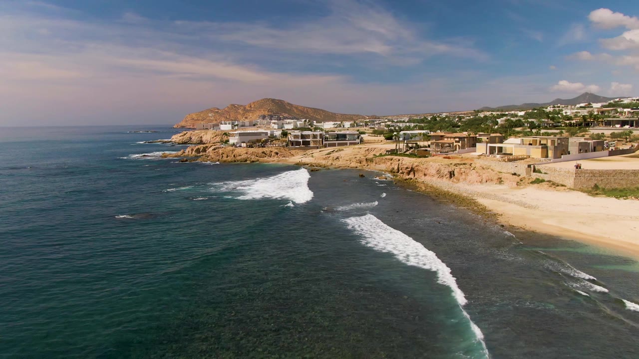 Wide aerial shoreline with a breaking wave, sandy beach, rocky point, and coastal development.