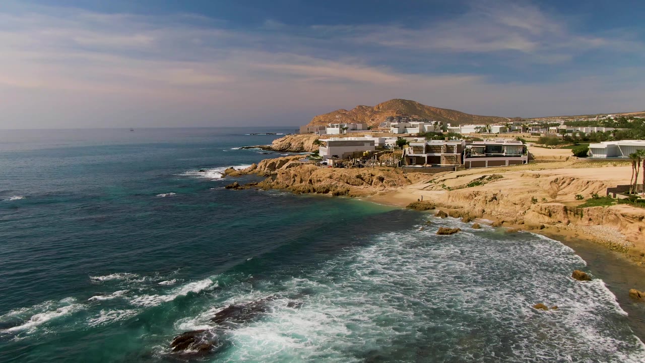 Elevated view over El Tule Los Cabos water where white surf spreads around dark rocks, with a tan sandy beach and coastal buildings beyond, no visible people.