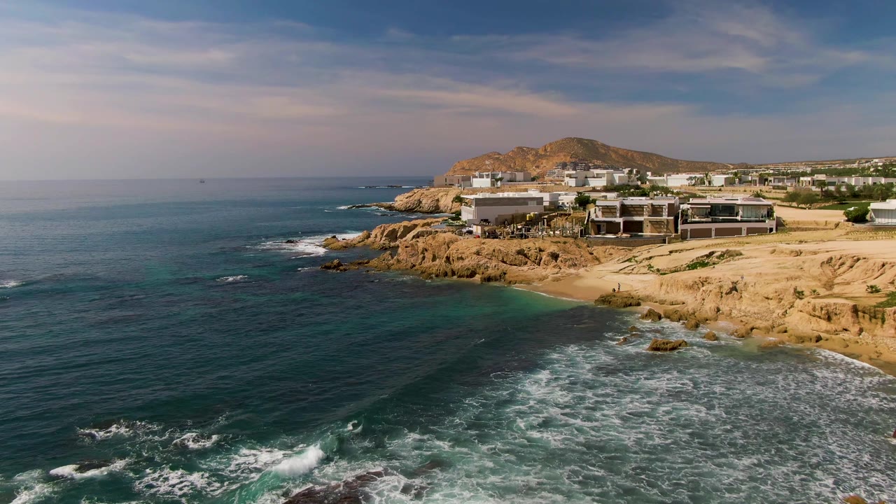 Aerial frame at El Tule Los Cabos with turquoise water near shore, foam curling around rocks, a small beach, and low coastal buildings under a clouded sky, no visible people.