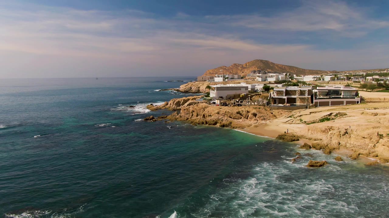 Wide aerial view in El Tule Los Cabos with more open water at left, foamy surf at lower right, rocky tan shoreline, beach, and coastal buildings, no visible people.