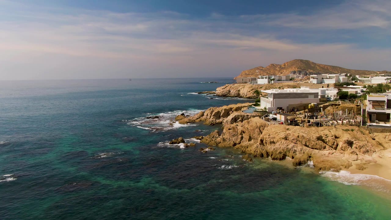 Aerial frame from El Tule Los Cabos showing turquoise water in the foreground, tan cliffs and white coastal buildings to the right, a small sandy beach below, and no visible people.