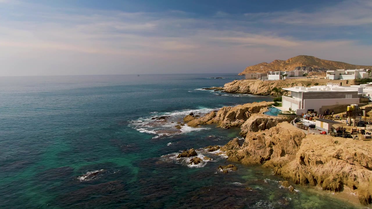 Aerial frame from El Tule Los Cabos showing the rocky shoreline closer to the right edge, white modern buildings above the rocks, turquoise water near shore, and no visible people.