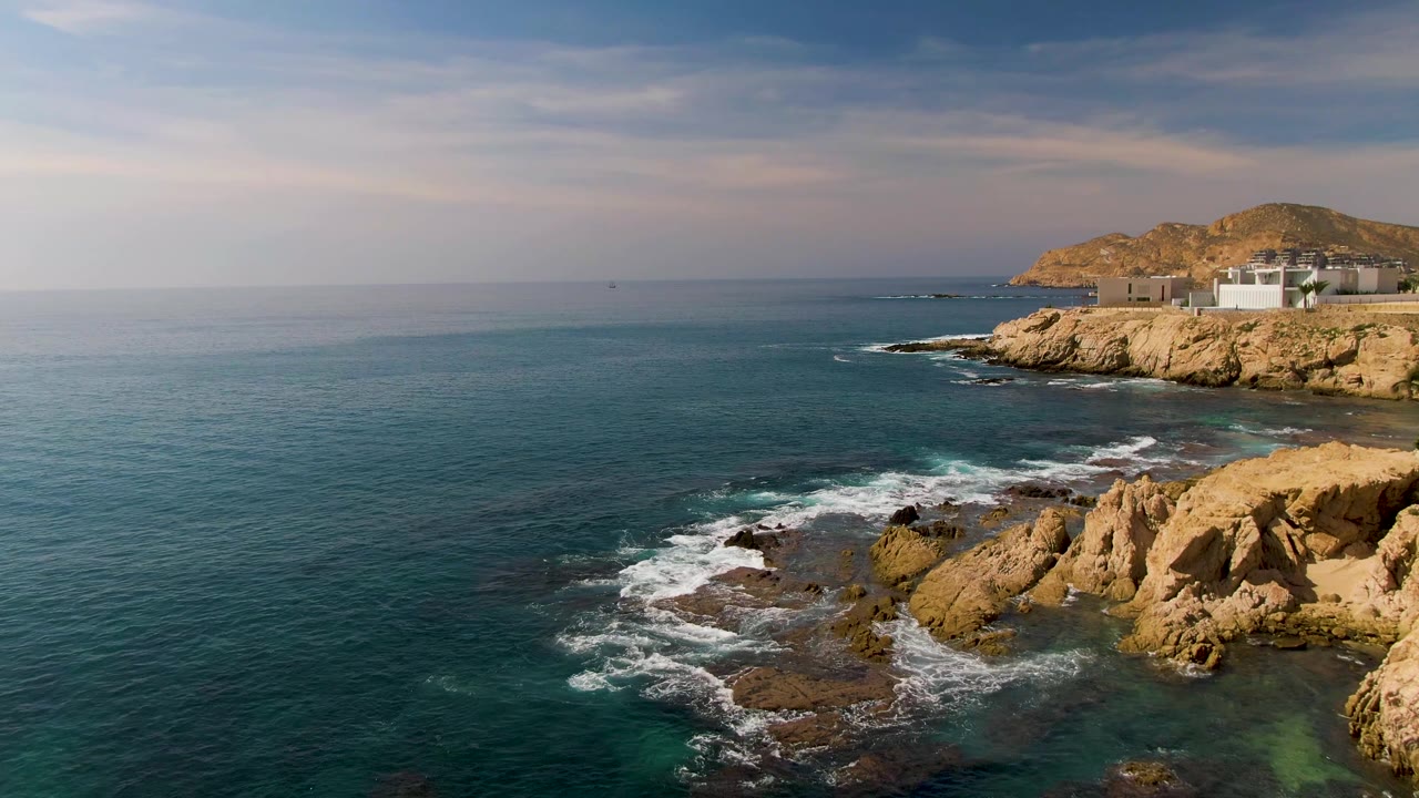 At El Tule Los Cabos, blue-green water rolls toward tan shoreline rocks with white foam in the lower frame, a rocky headland and low buildings at right, and no visible people.