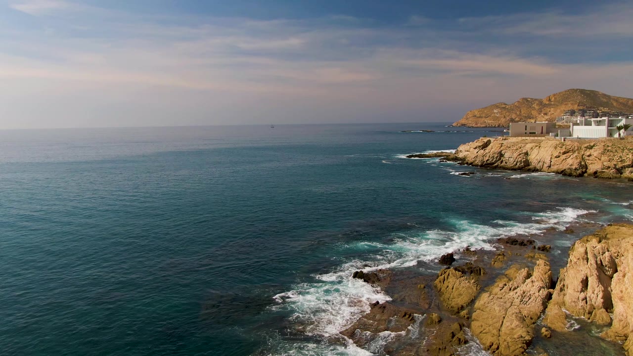 At El Tule Los Cabos, the wave foam pulls back from the dark rocks near the bottom edge, leaving calmer blue-green water across the center and a rocky headland at upper right, with no visible people.