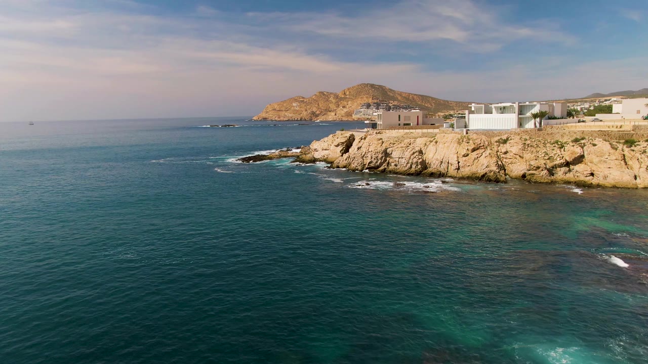 Elevated frame from El Tule Los Cabos shows deep blue ocean in the foreground, rocky cliffside buildings on the right, a low hill beyond the shore, and no visible people.