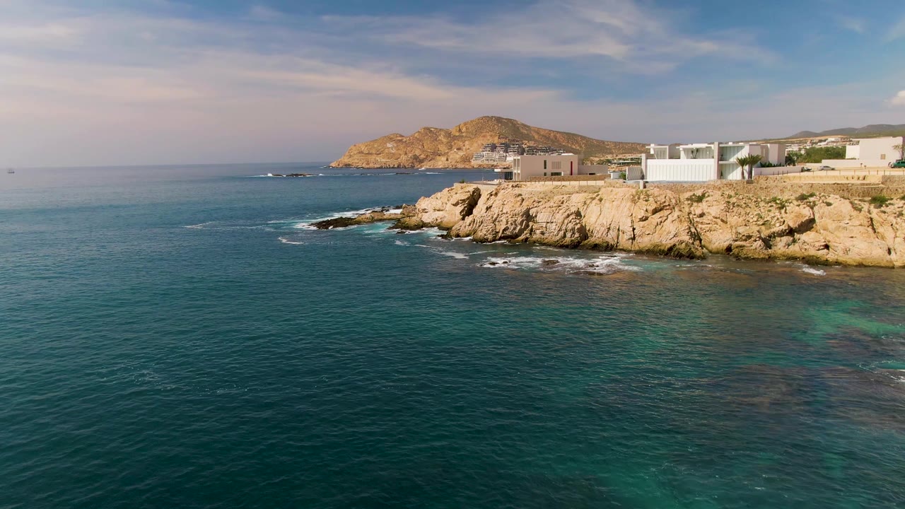 At 0:01 in El Tule Los Cabos, the ocean surface fills most of the frame while pale rocks, white buildings, distant hills, and no visible people remain along the right edge.