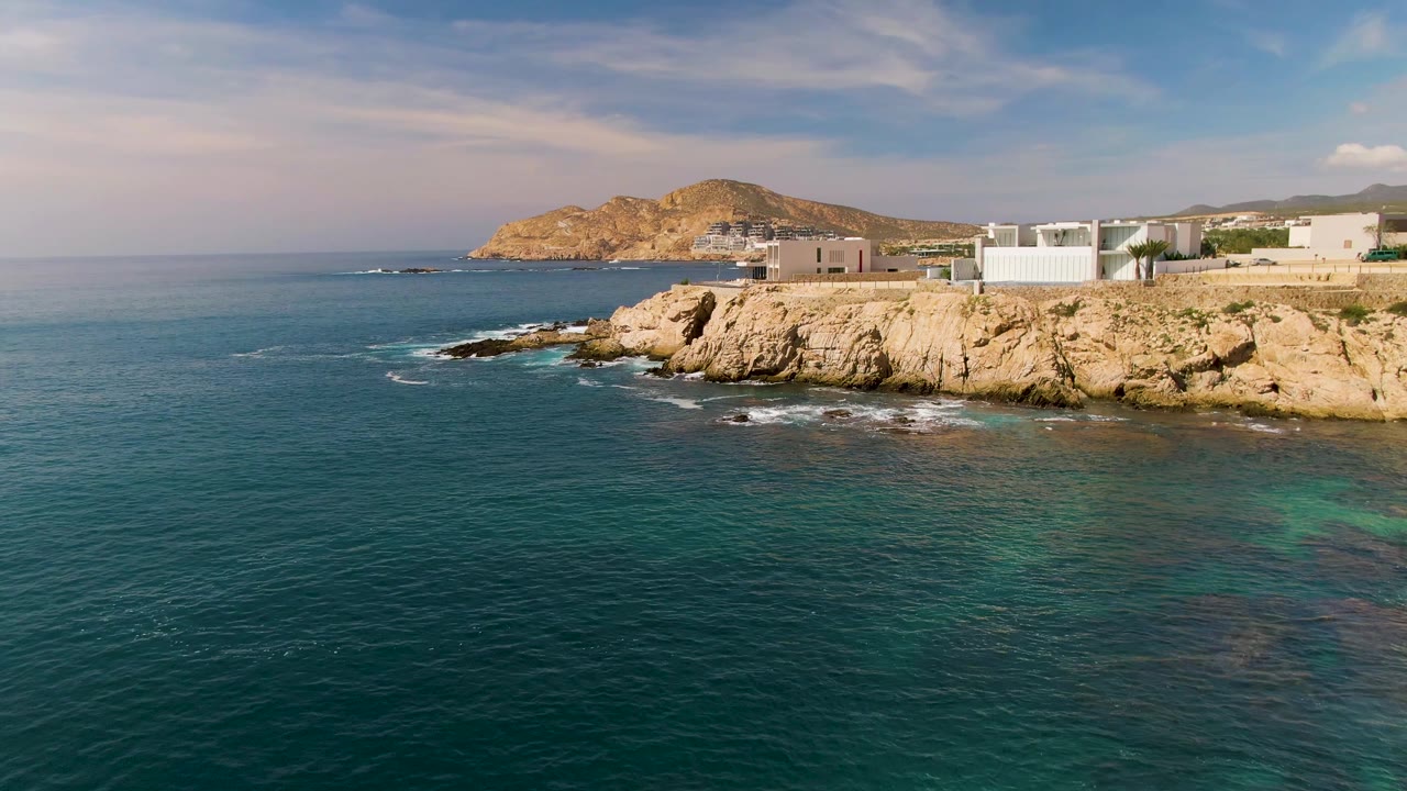 At 0:02, El Tule Los Cabos coastline runs across the frame with small whitecaps at the rocks, turquoise patches in the water, white structures above the cliff, and no visible people.