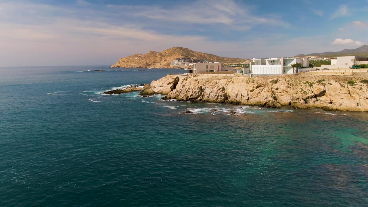 Wide coastal view with ocean foreground, rocks, buildings, and hills.