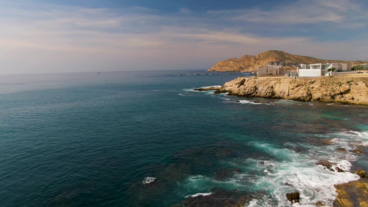 Opening frame of El Tule Los Cabos, Mexico, showing deep blue ocean, white foam over dark shoreline rocks, tan cliffs, low buildings, and no visible people.