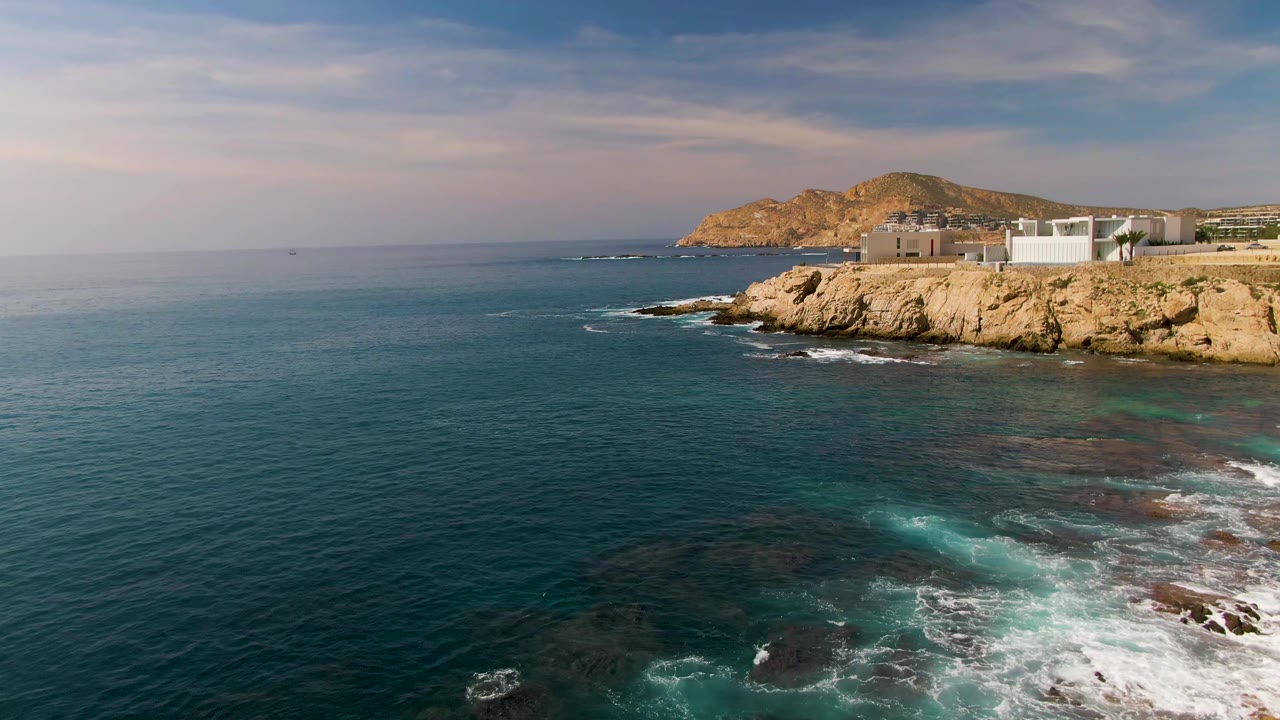 Second frame of El Tule Los Cabos, Mexico, with turquoise patches in the water, receding foam near the rocks, tan cliffs, low coastal buildings, and no visible people.