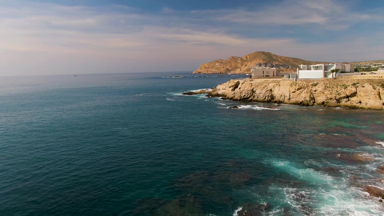 Third frame of El Tule Los Cabos, Mexico, where the open ocean occupies more of the frame, with a rocky point, cliffside buildings, thin surf lines, and no visible people.