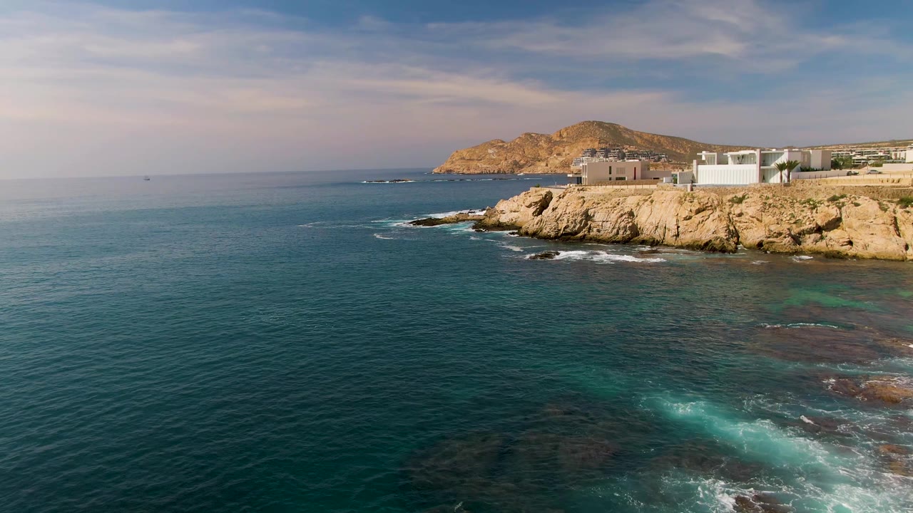 Fourth frame of El Tule Los Cabos, Mexico, showing calmer blue water in the foreground, small foam lines near the rocky cliff, low buildings above the shore, and no visible people.