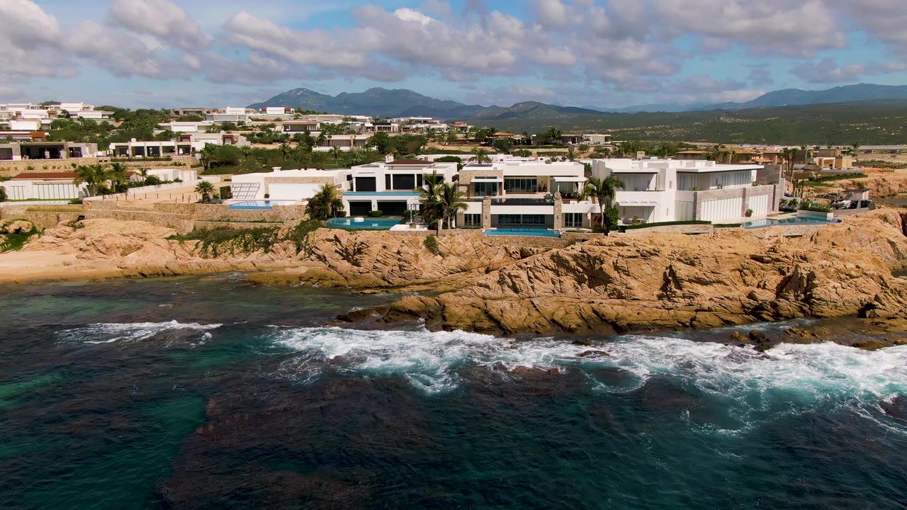 Opening frame of El Tule Los Cabos showing white modern buildings on tan rocks above dark blue ocean surf, with inland hills and scattered clouds behind them and no visible people.