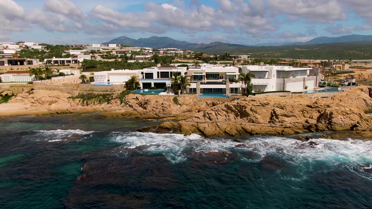 El Tule Los Cabos aerial frame with the modern buildings still prominent, waves spreading white foam over the rocks in front, green-blue water at lower left, and no visible people.