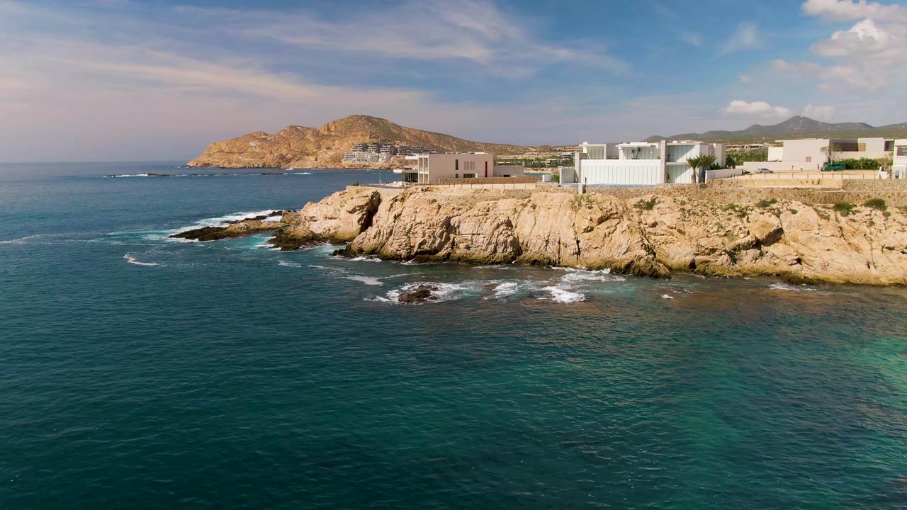 Aerial view over El Tule Los Cabos showing blue-green ocean, tan rocky cliffs, low coastal buildings, distant hills, scattered clouds, and no visible people at the opening moment.