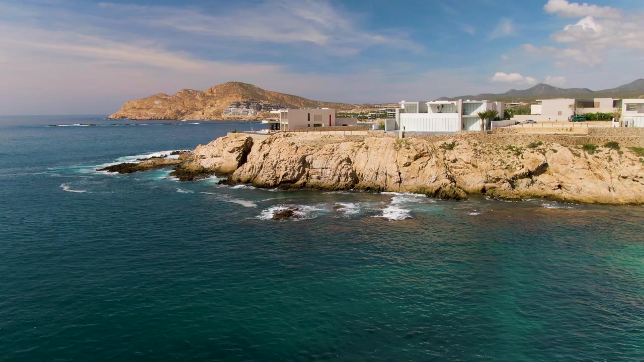 One second into the El Tule Los Cabos clip, waves leave white foam below the tan cliff while teal water fills the foreground and white buildings line the upper shore with no visible people.